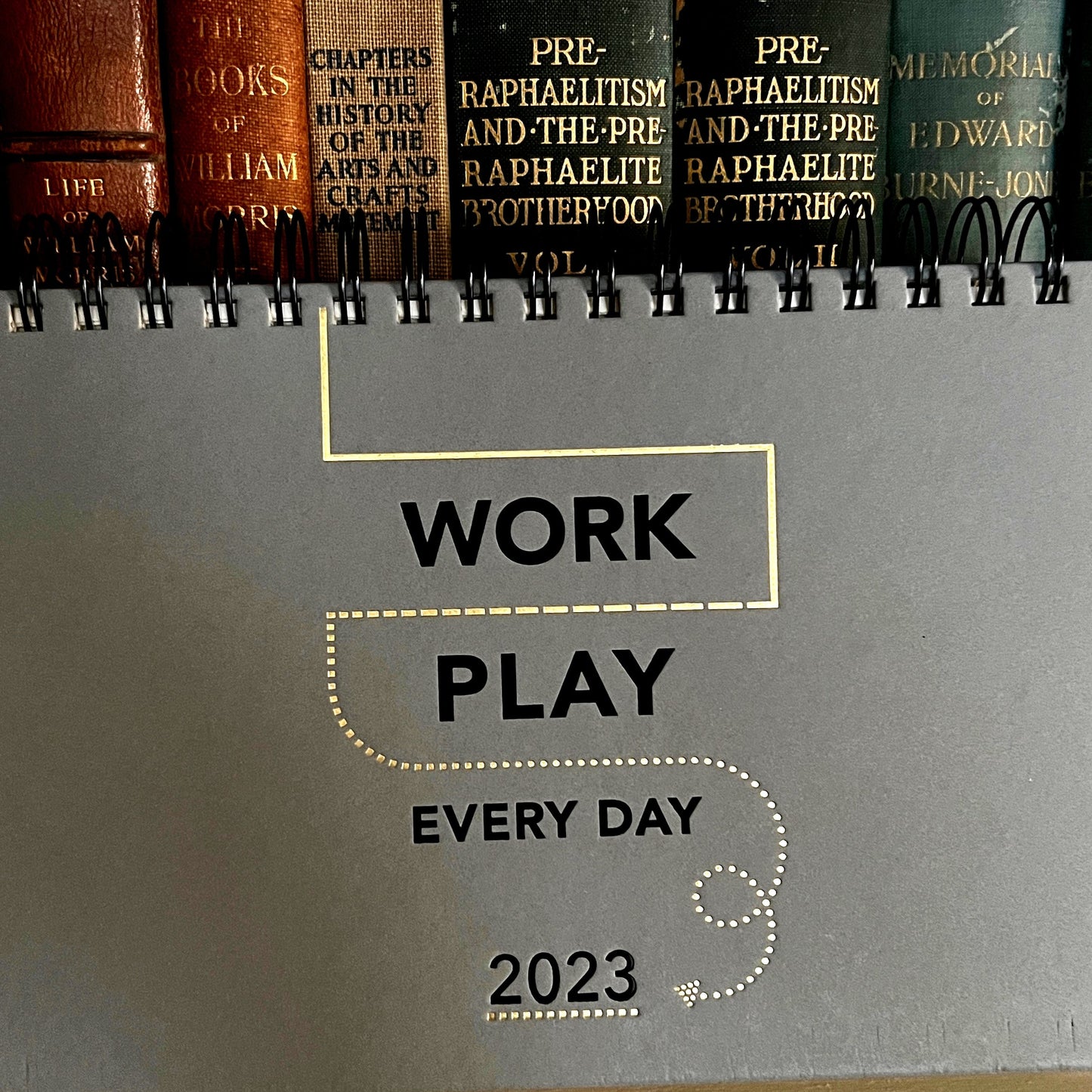 work play every day 2023 planner pictured in front of a row of antique gilded books about pre-raphaelitism. The planner has a slate grey hard cover and black wire-o spiral binding. The front has a foil-debossed design in champagne and black, with the words WORK PLAY EVERY DAY 2023 and a linear/dotted design